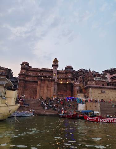 Populated ghat area near a historic structure at dusk.
