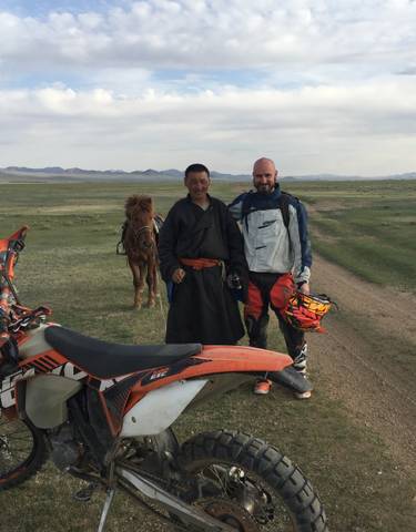 Two people standing beside motorcycles on a vast plain with a horse in the background.