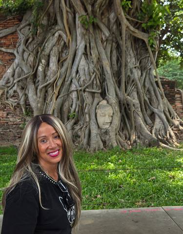 A woman sitting by a Buddha head embedded in a tree.