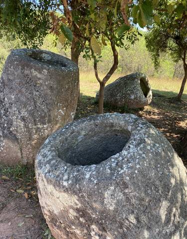 Stone jars on the ground surrounded by grass and trees.