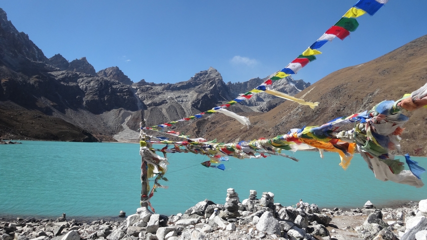 Colorful prayer flags hanging over a turquoise lake with mountains in the background.