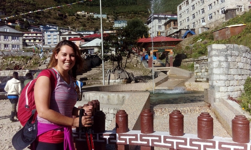 A person smiling in front of traditional Nepali architecture with other people in the background.