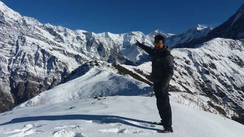 Hiker points toward a jagged snow-covered Himalayan ridge while standing on a white snowy slope under a deep blue sky.