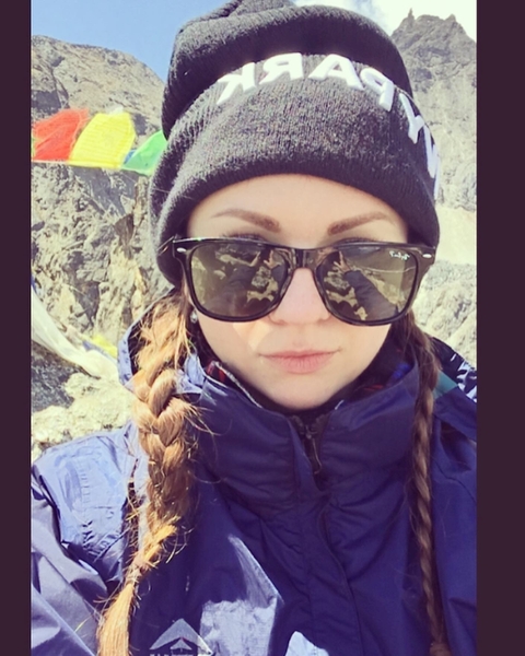 Selfie of female trekker wearing sunglasses and winter hat in a rugged mountain setting.