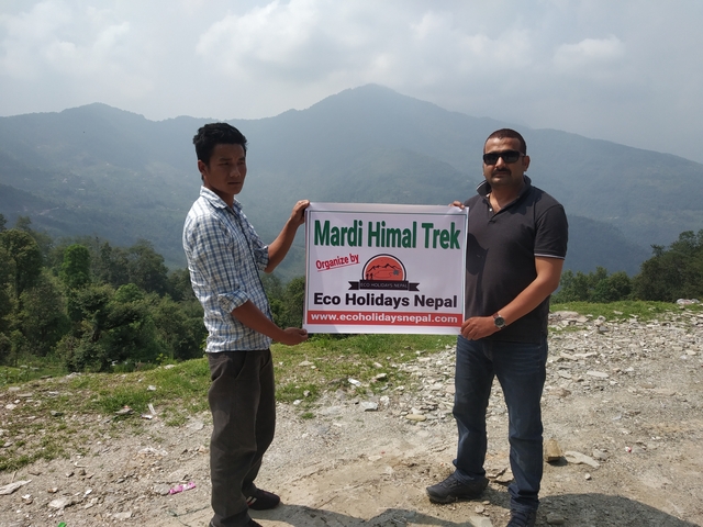 Two men hold a banner advertising a Mardi Himal trek against a hazy Himalayan backdrop.