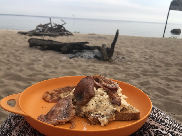 A plate of breakfast food on a beach with a bonfire setup.