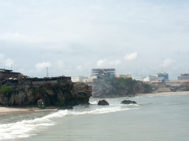 Hazy coastal view of rocky cliffs, crashing waves and urban buildings in the distance.