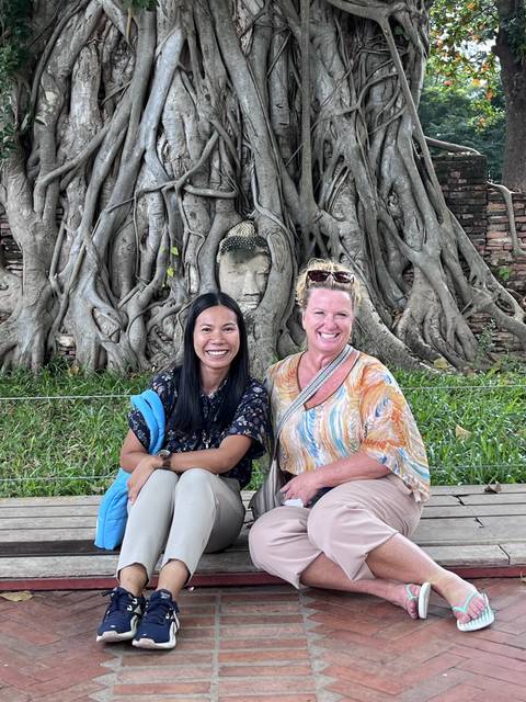 Two women sitting next to a tree with a Buddha head intertwined in roots.