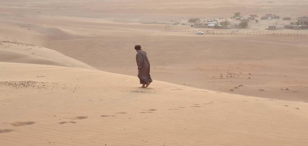 A person walking barefoot across sand dunes with scattered settlements.
