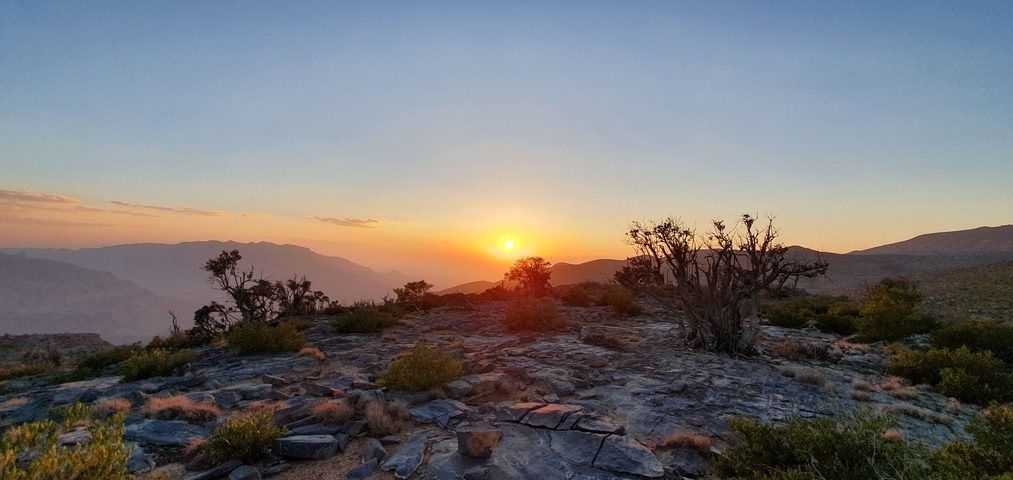 A sunset over rocky terrain with shrubbery and distant mountains.