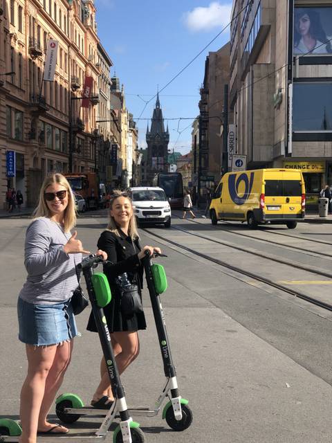 Two people posing with scooters on a street with vehicles and buildings in the background.