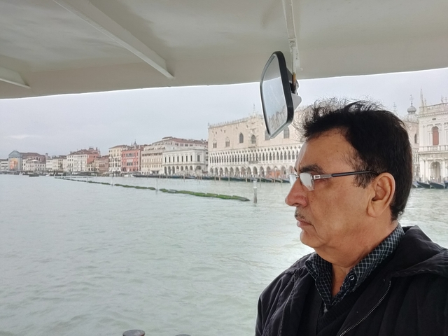 Man on a boat with a view of city buildings on a cloudy day.