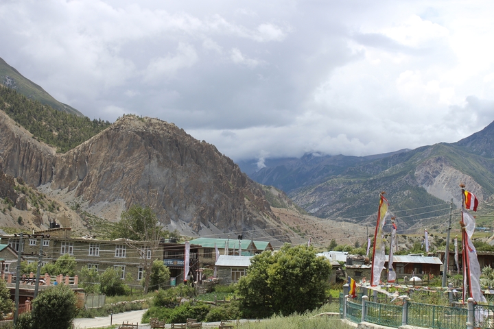 Mountain village beneath dramatic cliffs under a cloudy Himalayan sky.