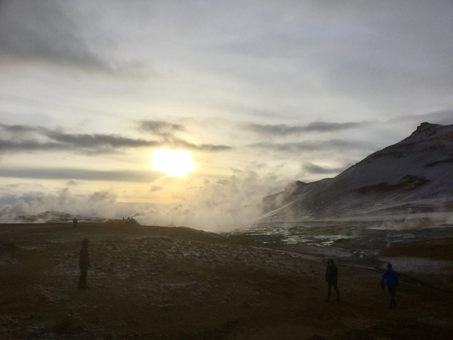 Sun partially hidden by clouds over a geothermal area with people walking.
