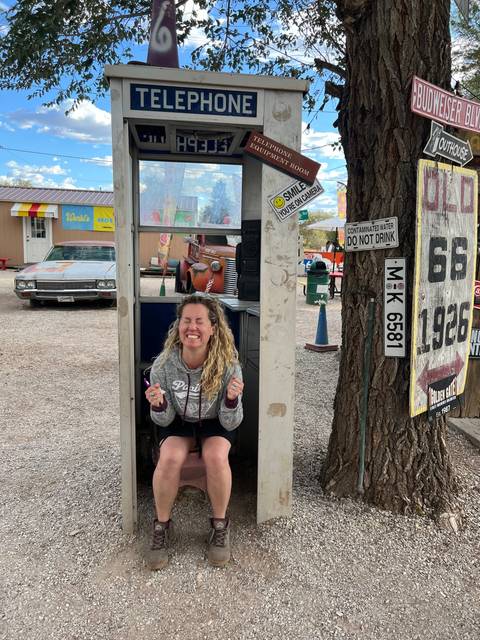 A person sitting inside a retro telephone booth surrounded by signs and trees.