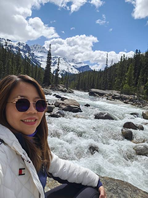 Woman at river with rapids, trees, and mountains.