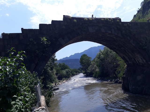 Stone bridge arching over a river with mountains in the background.