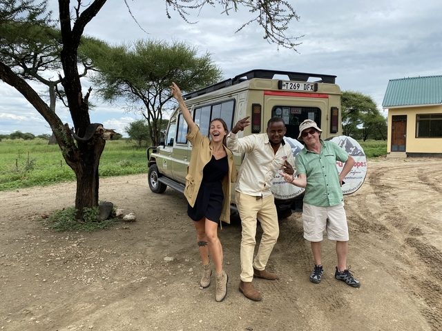 Guide and tourists celebrate beside safari vehicle under acacia tree