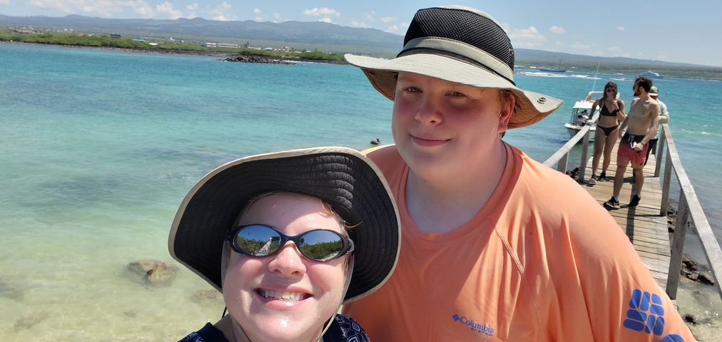 Two people taking a selfie on a boardwalk with the sea and mountains in the background.