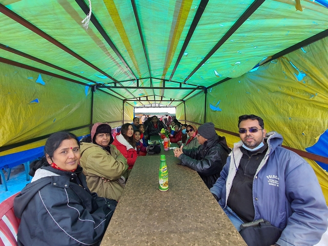 Large group of people sitting under a colorful tarp.