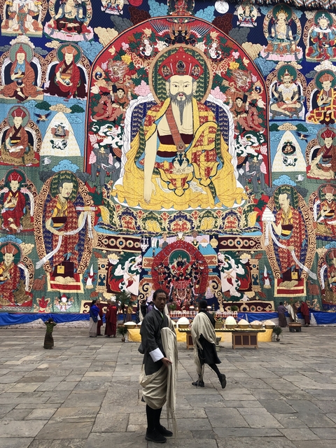 Colorful religious tapestry with elaborate Buddhist figures displayed during a festival, visitors gathered below.