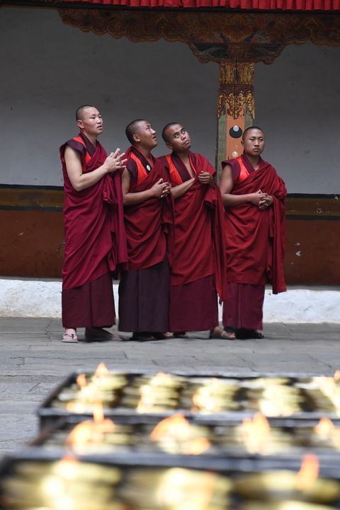 Four young monks in deep red robes stand in line with folded hands against a monastery wall.