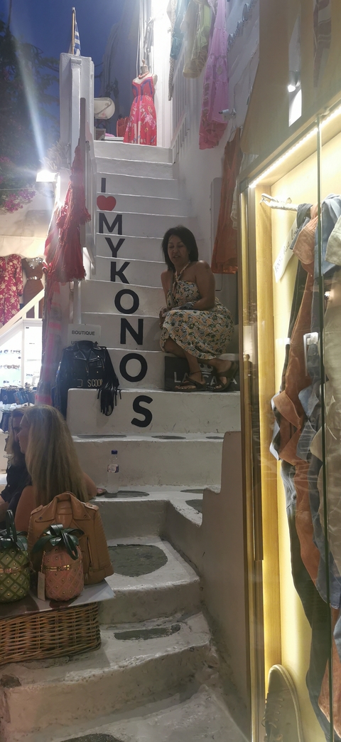 Woman in patterned dress poses on narrow white stairs in a bustling Mykonos alleyway.