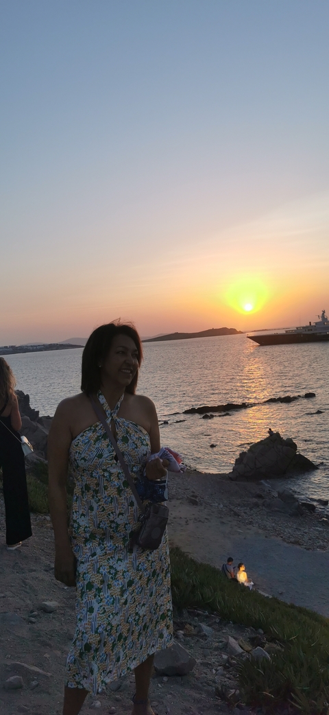 Woman smiles in front of a vivid Aegean sunset and calm sea with distant islands.
