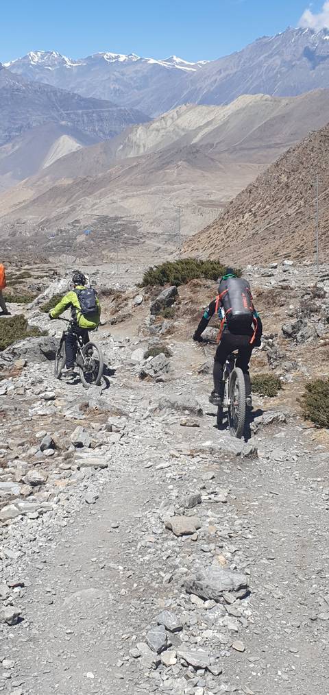 Two mountain bikers on a rocky path with mountains in the background.