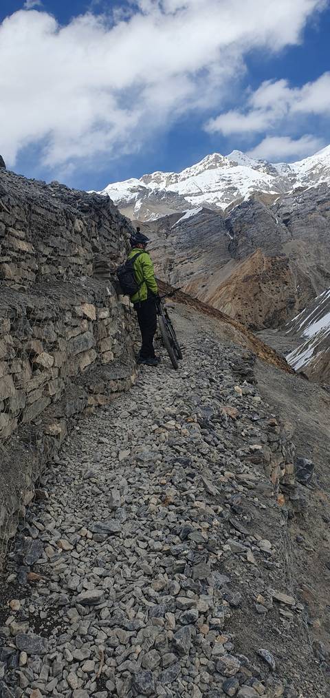 Biker resting on a rocky mountain path with snowy peaks visible.