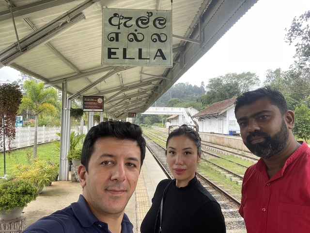 Three travellers take a selfie under the ELLA train station sign beside railway tracks