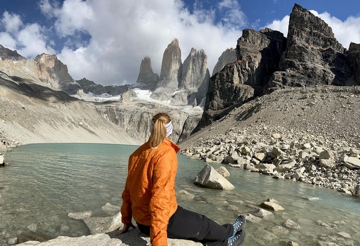 Traveler in an orange jacket sits on a rock looking across the turquoise lake toward the Torres del Paine towers