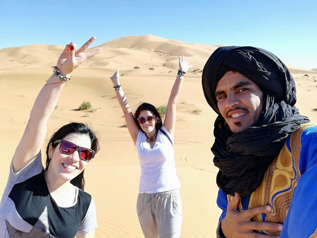 Three smiling travellers and a turbaned local guide pose joyfully in golden Saharan dunes.