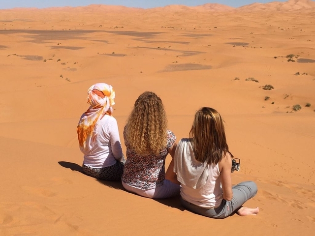 Three women sit on a sand dune looking out over endless rolling Sahara desert.
