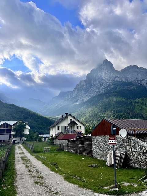 Alpine village houses beneath towering jagged peaks with dramatic sunlit clouds in the Accursed Mountains.