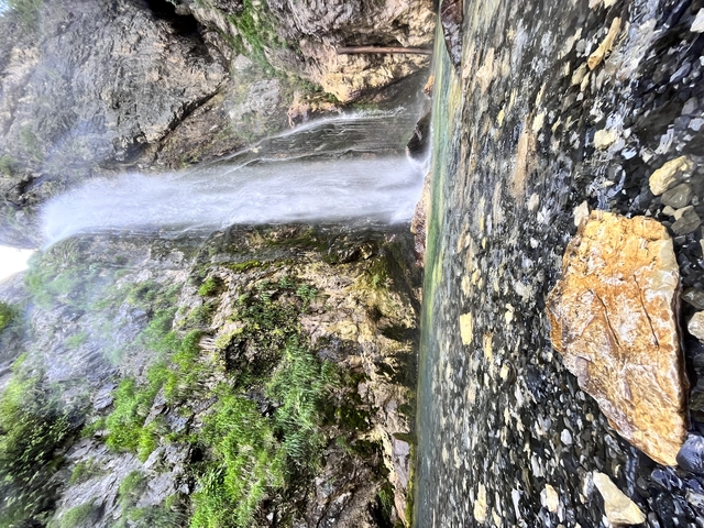 A powerful waterfall plunges down mossy rock walls beside a stony riverbank.