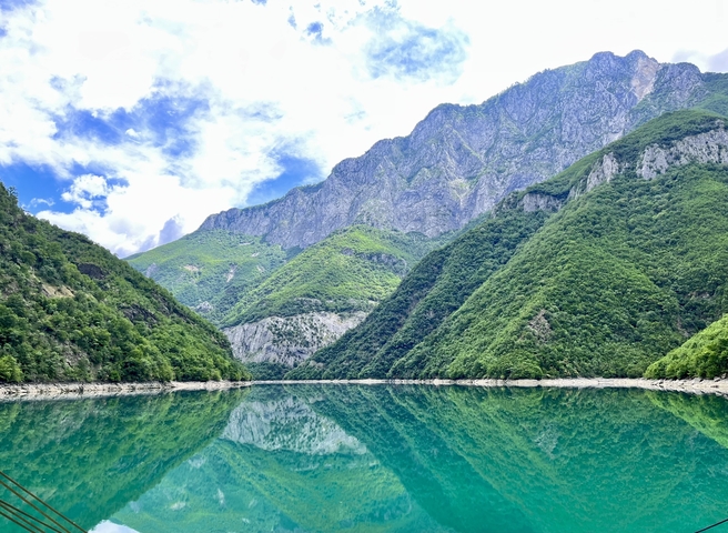 Crystal-clear turquoise lake perfectly reflects steep green mountains and cliffs under a bright sky.