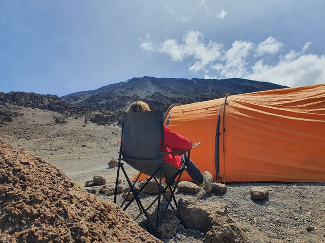 Person sitting in front of a bright orange tent in a rocky, mountainous area.