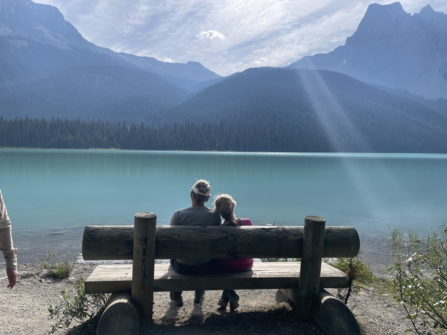 Two people sitting on a bench overlooking a calm lake with mountains in the distance.
