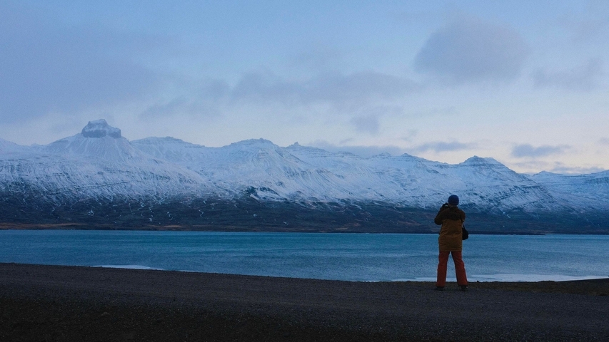 A person standing by a large lake with snowy mountains in the background.