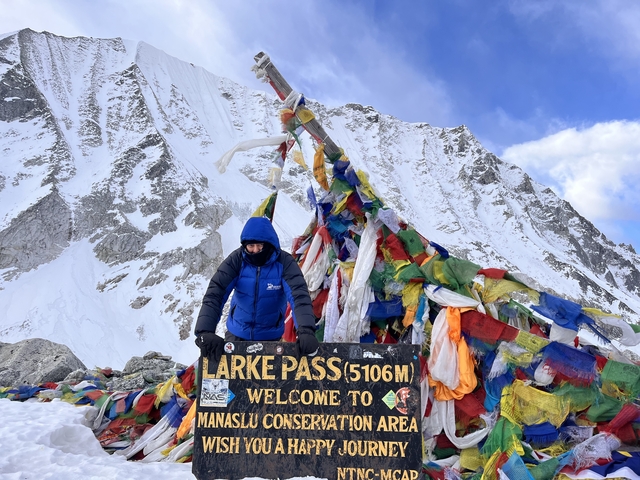 Person at Larke Pass sign with colorful prayer flags and snowy mountain background.