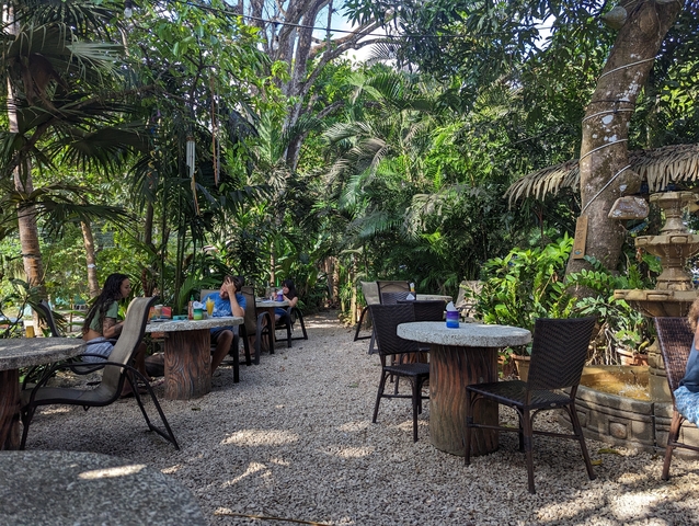 Outdoor cafe with tables and chairs, surrounded by greenery.
