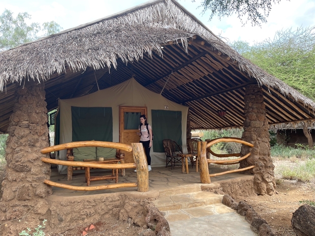 Person standing on a porch of a hut in a camp-like setting.
