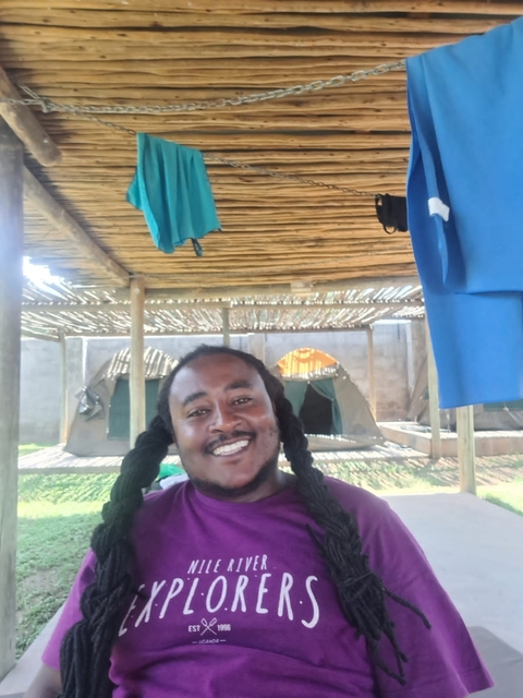Person smiling under a wooden structure with tents in the background.
