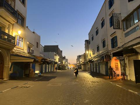 A street in Essaouira at dawn with lights on and a few people.