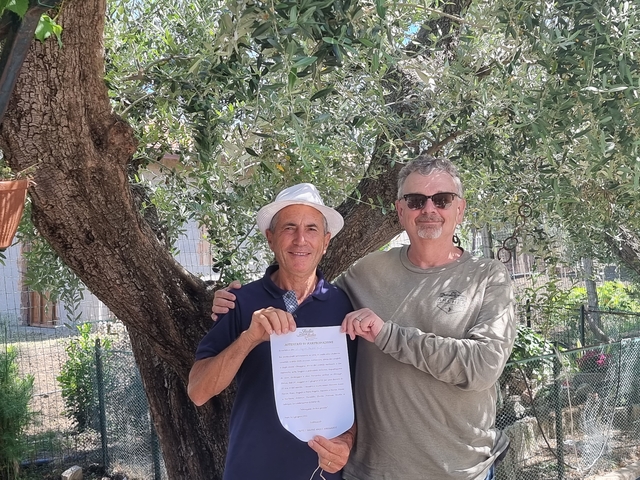 Two men holding a certificate under an olive tree.