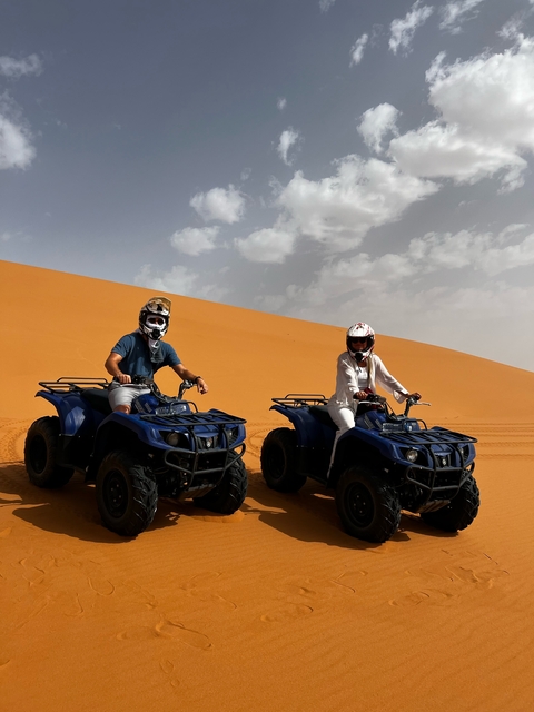 Two people on quad bikes in the desert with dunes in the background.