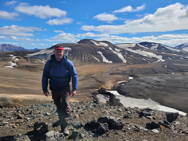 Person hiking on volcanic terrain with snow patches.