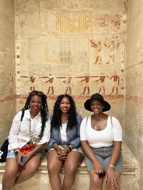 Three people smiling indoors with ancient wall carvings.