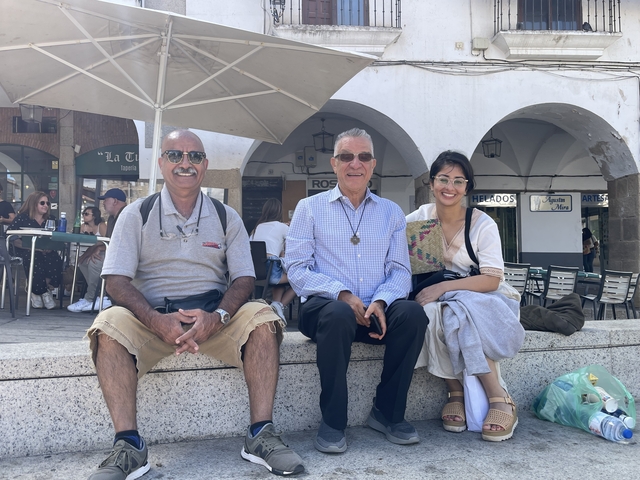 Three people sitting under an umbrella in a plaza.